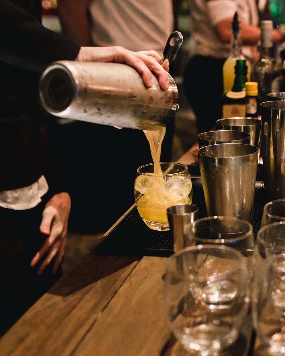 Barkeeper gießt einen Drink aus dem Shaker, daneben Gläser auf der Holztheke